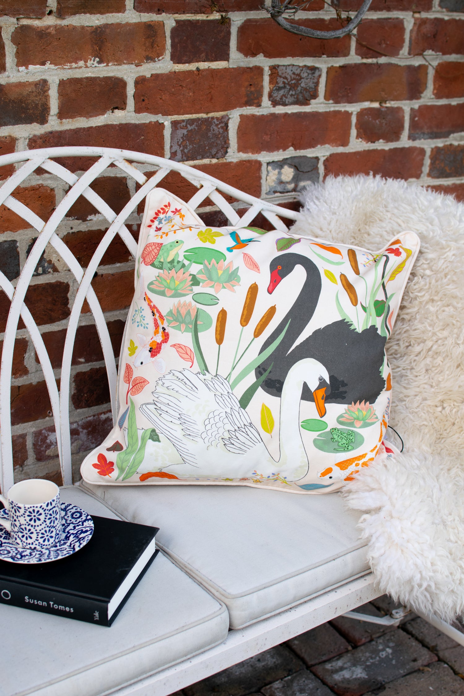 Karen Mabon Swan Lake Cushion with cream base featuring one black and one white swan, among reeds, lily pads and leaves.