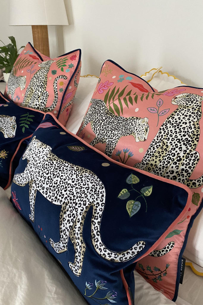 Two matching Leopard Bolster cushions on a bed with matching pink Snow Leopard cushions.Leopard Bolster Cushion, navy blue velvet with white snow leopard embroidery surrounded by leaf motifs, edged with pink contrast velvet trim. 